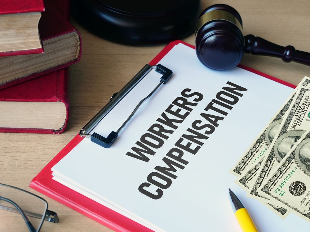 Clipboard labeled Workers Compensation with legal books, a gavel, and cash on a desk.