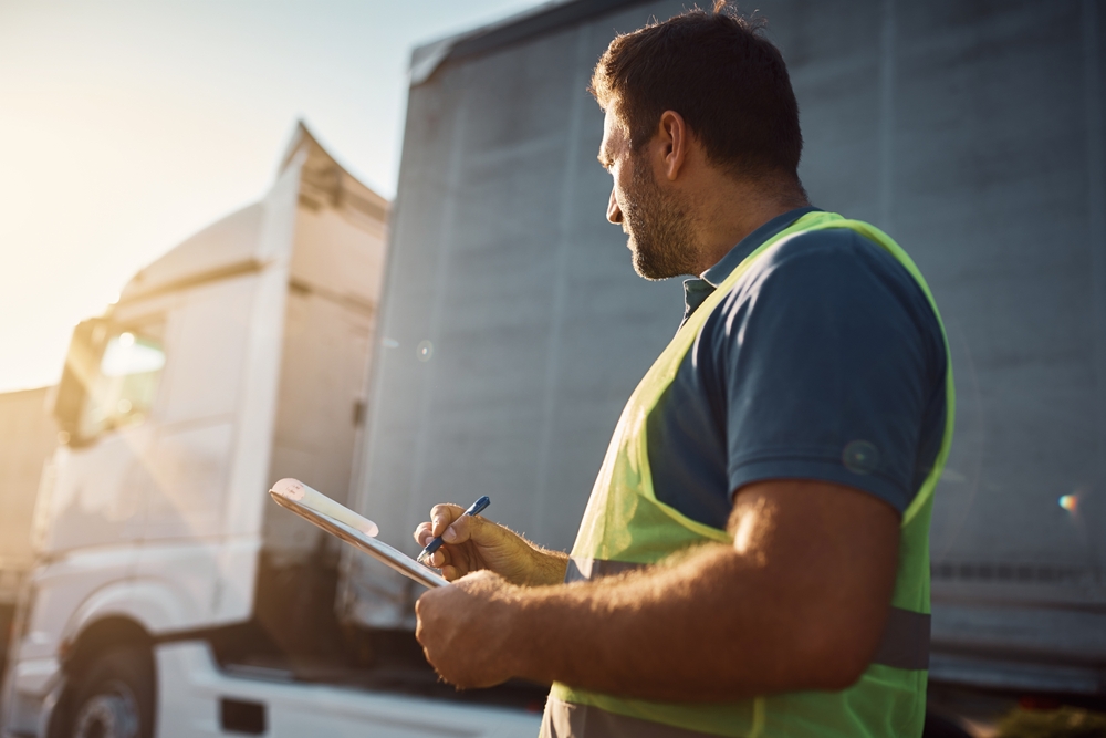 Truck driver performing a pre-trip inspection with a checklist beside a semi-truck.